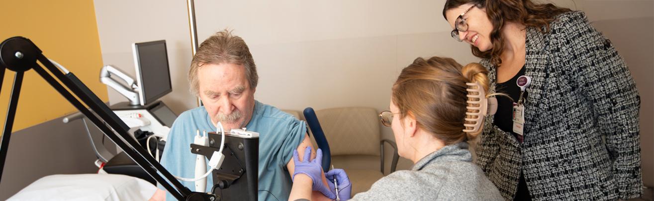 A fellow is adminitering an injection to a patient, under the guidance of an attending physician